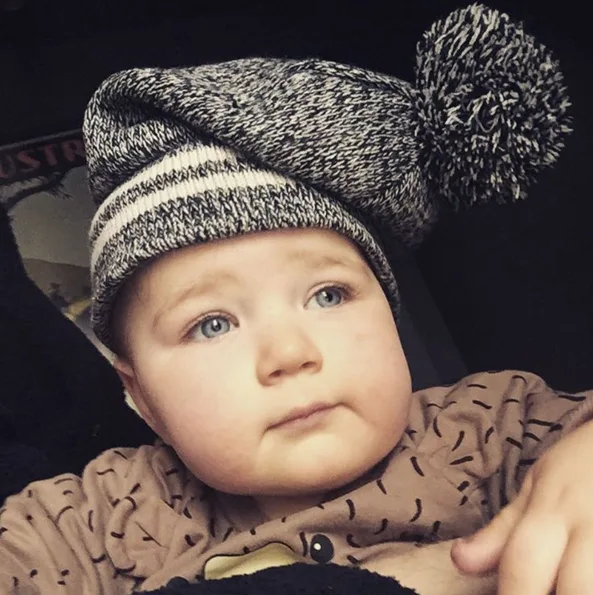 A baby wearing a gray and white knit hat with a pom-pom, looking upward with a neutral expression.