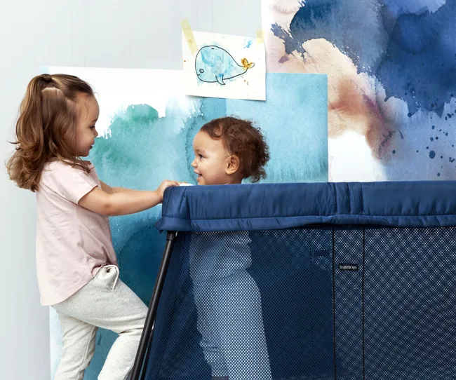 Two young children playing near a blue travel cot, with watercolor artwork in the background.