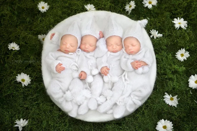 Four sleeping newborns in white outfits and hats lying on a soft surface surrounded by daisies.