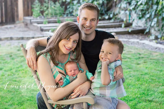 Family of four sitting together outdoors, parents smiling, holding a baby and a young child on a chair in a garden setting.