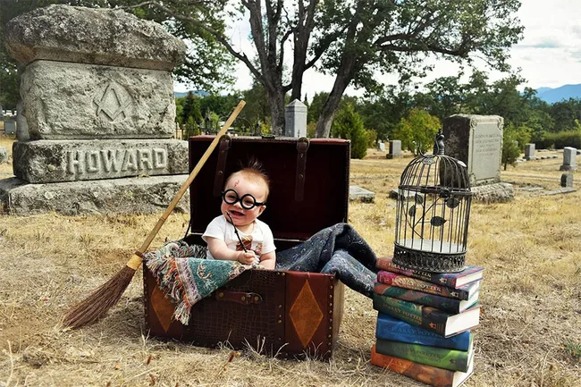 Baby dressed as Harry Potter sits in trunk with broom, books, and cage in a cemetery.