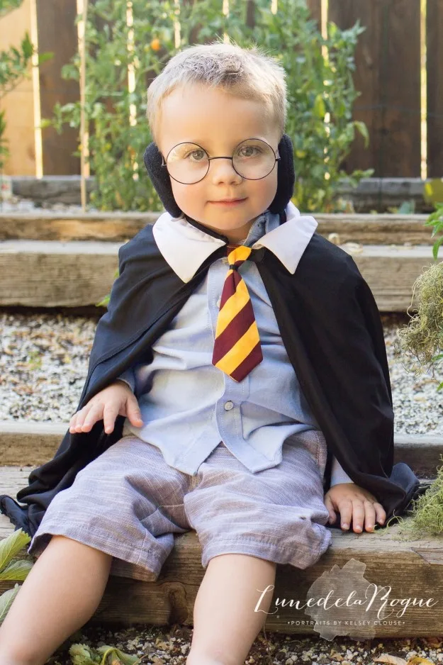 Toddler wearing glasses and a Gryffindor-themed cape and tie, sitting outside on wooden steps.