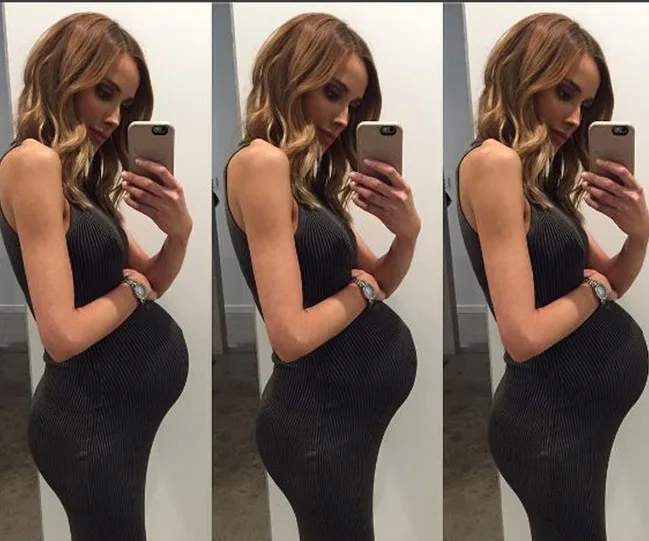 A pregnant woman taking a mirror selfie in a fitted dress, showing her side profile.