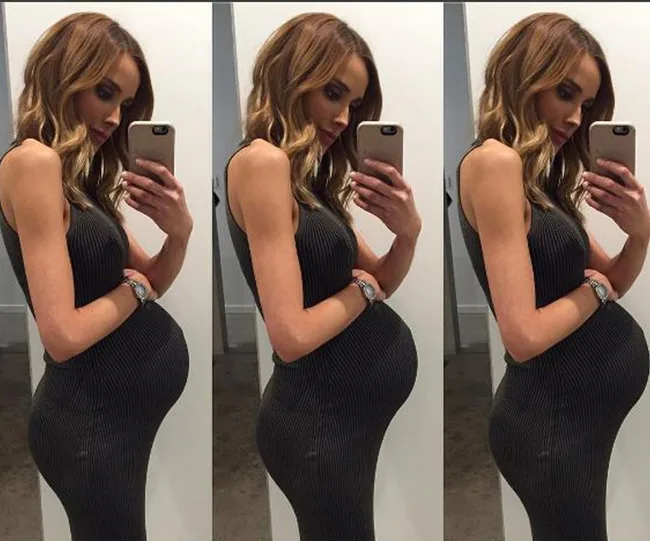 A pregnant woman taking a mirror selfie in a fitted dress, showing her side profile.