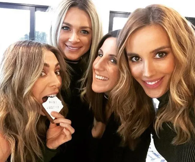 Four women smiling, one holding a cookie that says "Baby Juddy." They are posing closely together.