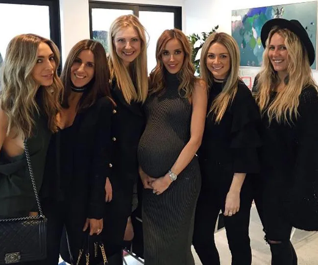 Group of six women posing indoors, one is visibly pregnant, all smiling at the camera.