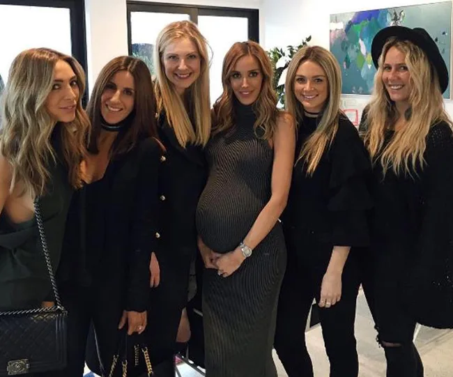 Group of six women posing indoors, one is visibly pregnant, all smiling at the camera.