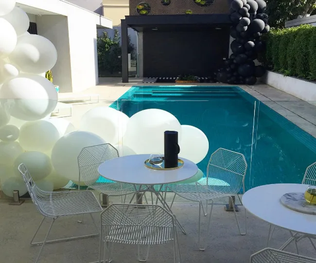 Outdoor pool area with white and black balloons, glass fence, and white tables and chairs.