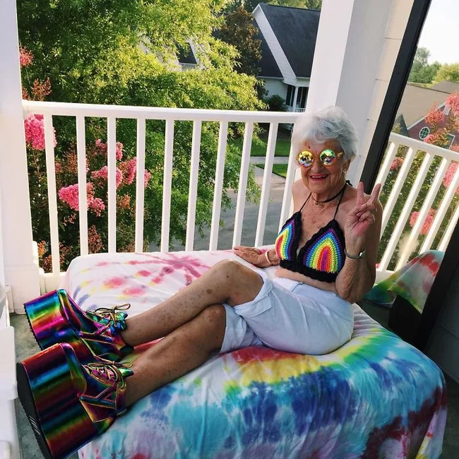 Elderly woman in rainbow outfit and glasses, posing with peace sign on a balcony with tie-dye bedding.