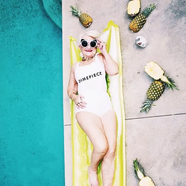 Elderly woman in a "Dimepiece" swimsuit lounges on an inflatable by a pool, surrounded by pineapples.