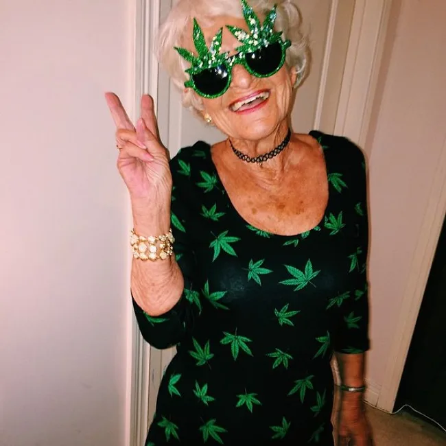 Elderly woman in a marijuana leaf dress and green leaf sunglasses making a peace sign gesture.