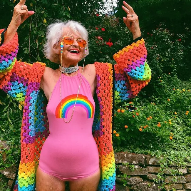 Elderly woman in rainbow outfit laughs joyfully with arms raised, standing in a garden.