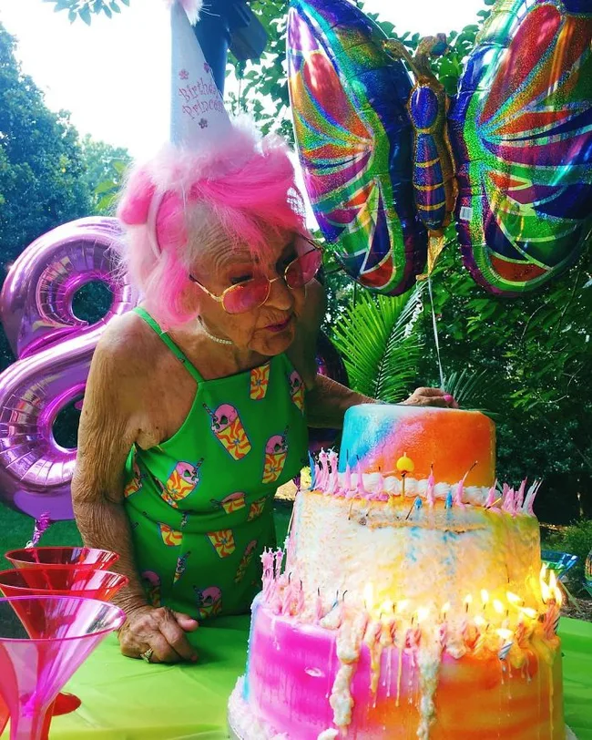 Elderly woman with pink hair blows out candles on a colorful birthday cake, wearing a party hat, with bright balloons.