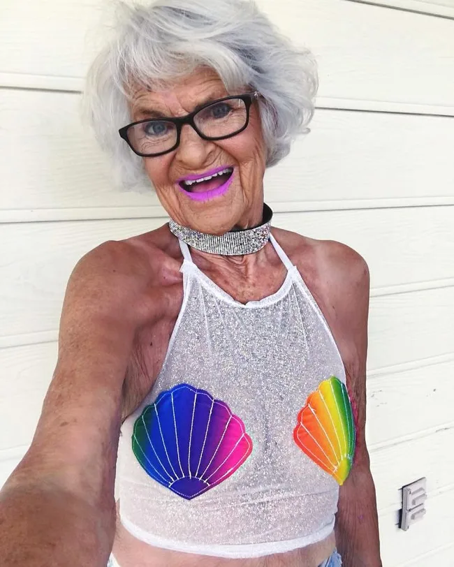 Older woman with gray hair and glasses, wearing a sparkly top with rainbow shell designs, smiling brightly.