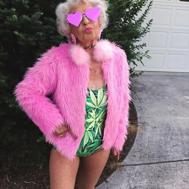 Elderly woman in a pink fur coat and heart-shaped glasses, wearing a green leaf-patterned swimsuit.