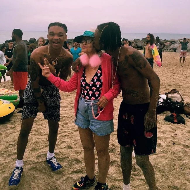 Three people on a beach, one kissing an older woman; all are smiling, with beachgoers in the background.