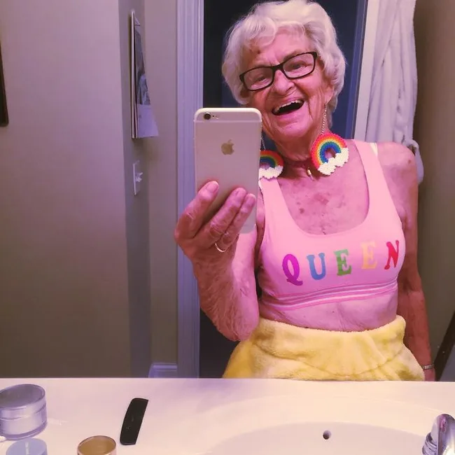 Elderly woman taking mirror selfie in "QUEEN" tank top with rainbow earrings, smiling in bathroom.