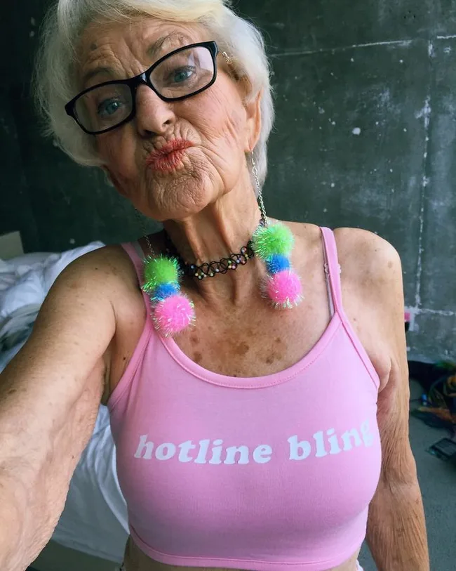 Elderly woman in glasses and pink "hotline bling" top with colorful pom-pom earrings, posing with a kissy face.