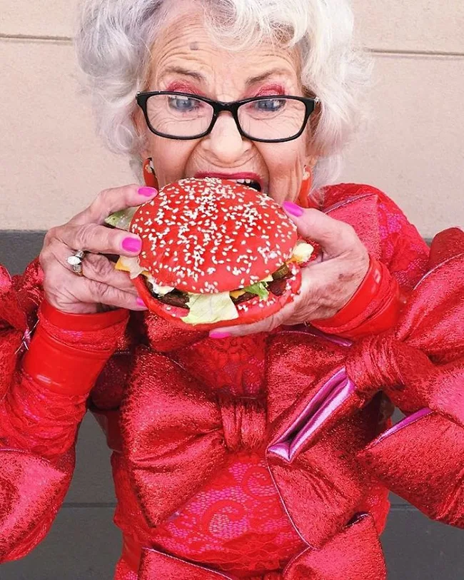 Elderly woman in red dress joyfully biting into a large red burger with sesame seed bun.