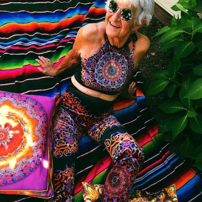 Elderly woman in vibrant, patterned outfit and sunglasses, sitting on a colorful striped blanket outdoors.