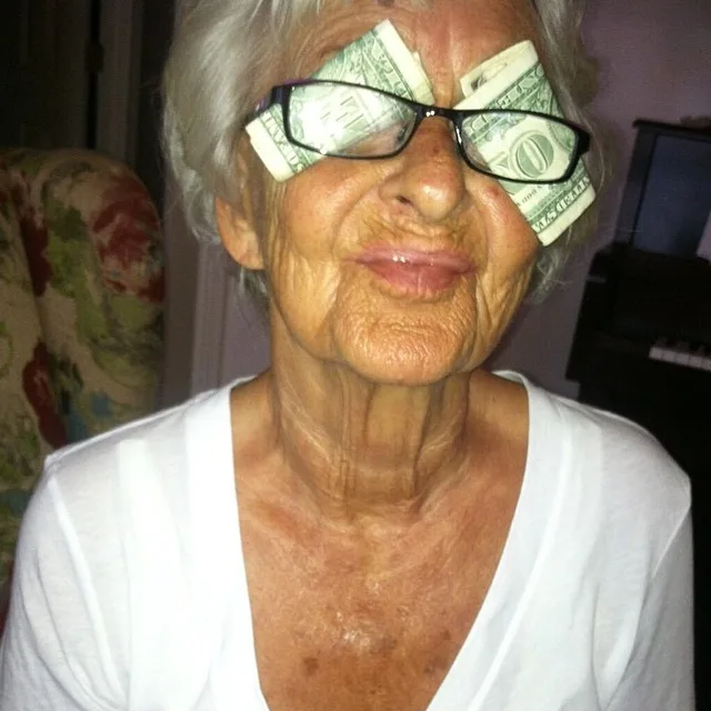 Elderly woman with dollar bills tucked in glasses, smiling with eyes closed, wearing a white shirt.