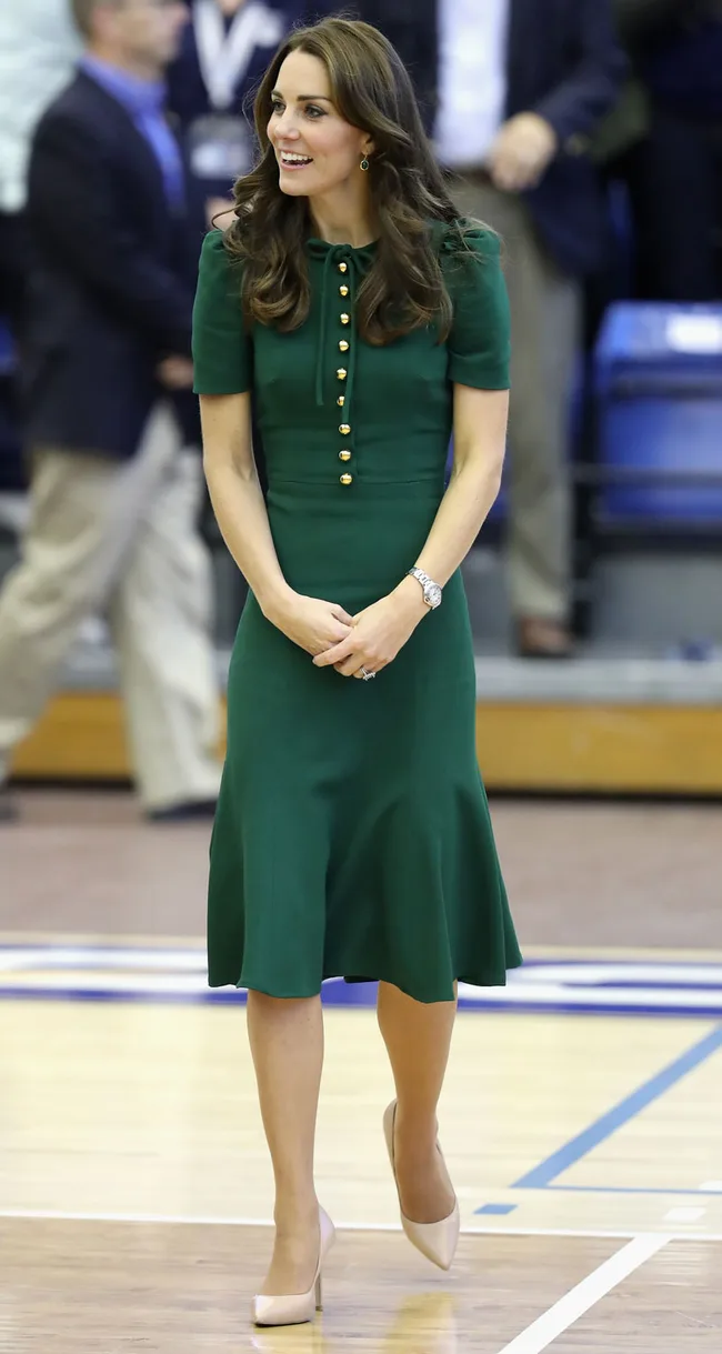 A woman in a green dress and beige heels stands on a gym floor, smiling, with people in the background.