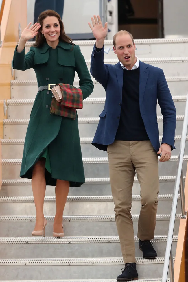 Couple waving while descending airplane stairs; woman in a green coat and man in a blue blazer.