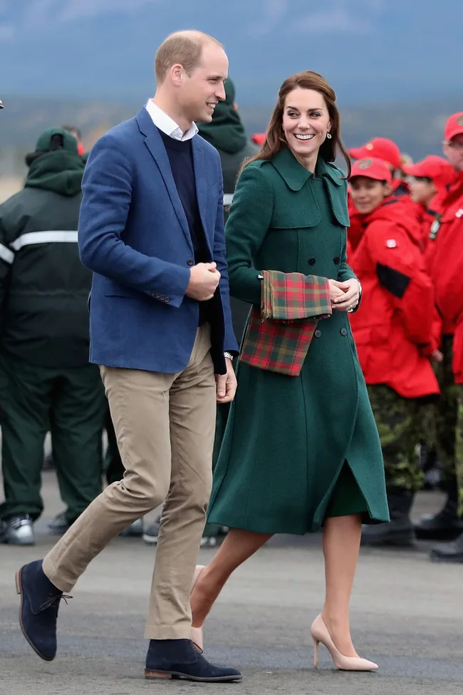 Elegant couple walking outdoors; woman in green coat with tartan scarf, man in blue blazer. People in red jackets background.