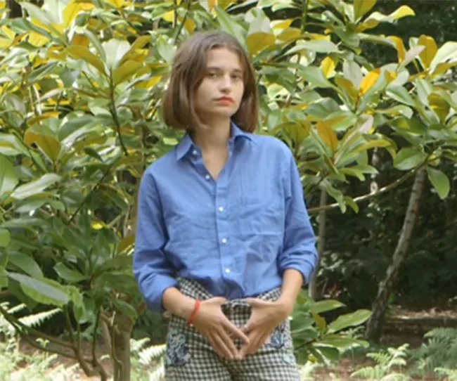 Young woman in a blue shirt and checkered pants standing in a garden with lush green foliage.