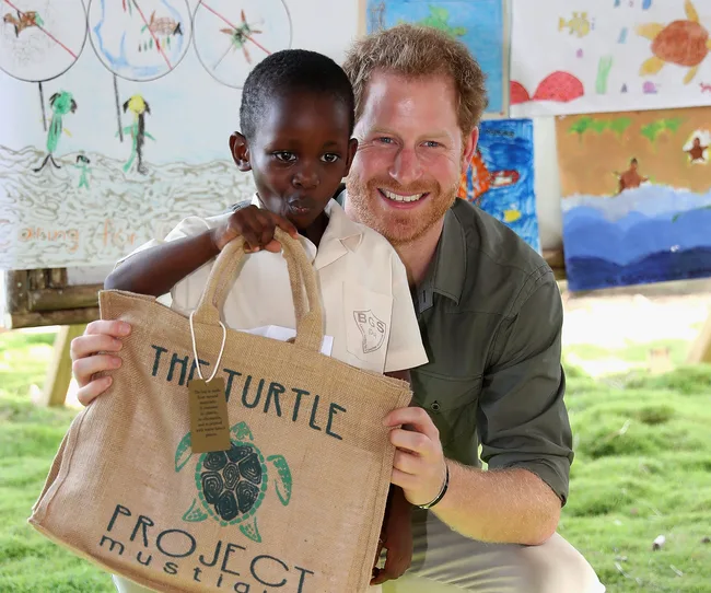 A man kneels next to a child holding a "The Turtle Project Mustique" bag, with colorful drawings in the background.