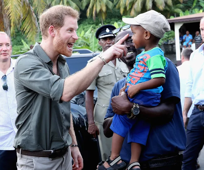 A man joyfully interacts with a child held by another man in a crowd with greenery in the background.