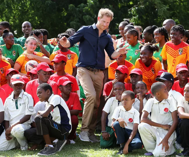 A person in a blue shirt interacts with a group of smiling children in sports uniforms and red caps outdoors.