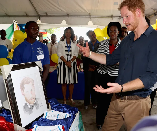 Man gestures near a portrait at a public event, surrounded by people and balloons under a tent.