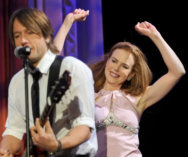 Keith Urban performs on stage while Nicole Kidman joyfully dances beside him.