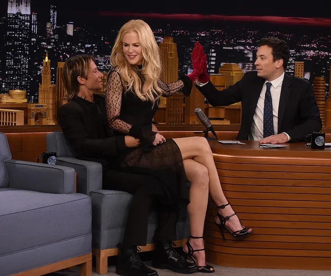 Celebrities on a talk show set, one seated and the other two engaging in conversation and gestures.