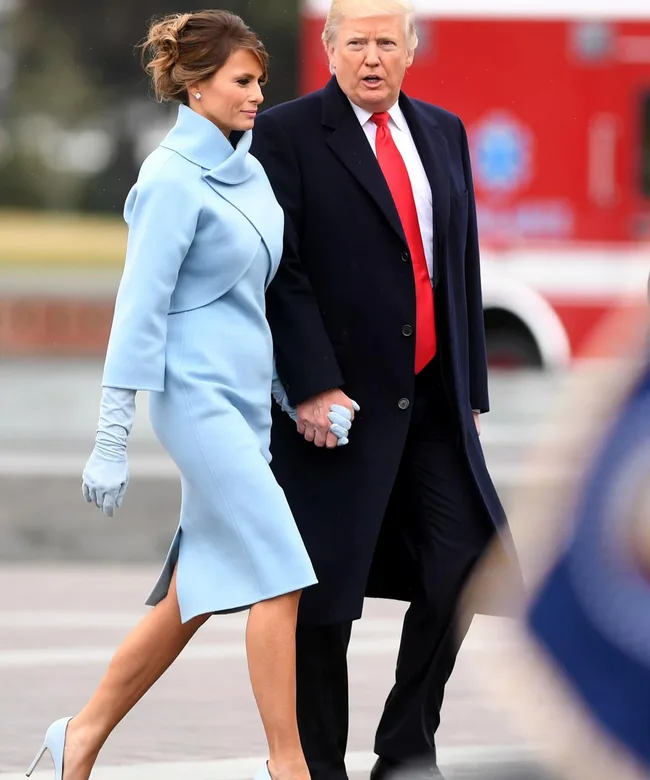 A man in a suit and red tie walks with a woman in a light blue coat and gloves, holding hands outdoors.