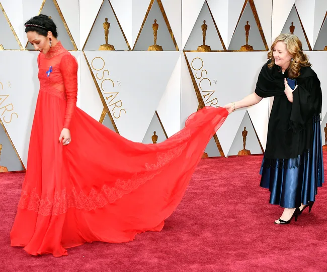 Woman in red gown walks red carpet at Oscars, another woman adjusts the dress train.
