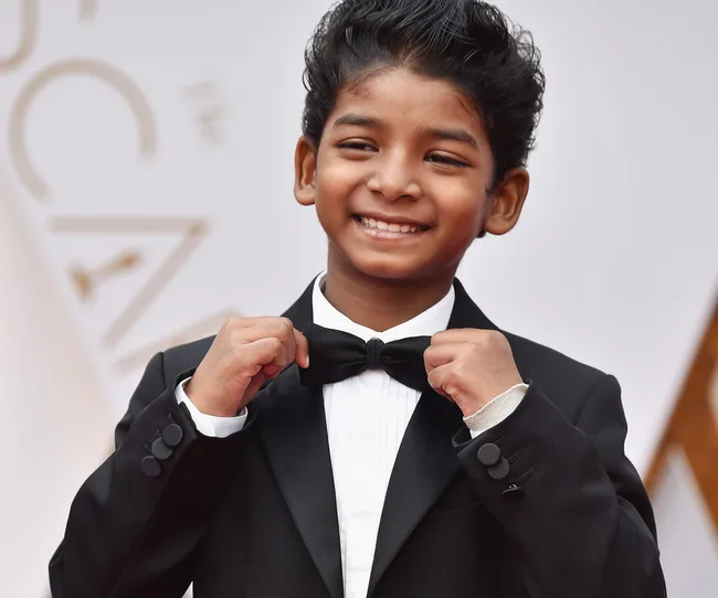 Young boy in a tuxedo smiling and adjusting his bow tie at an event.