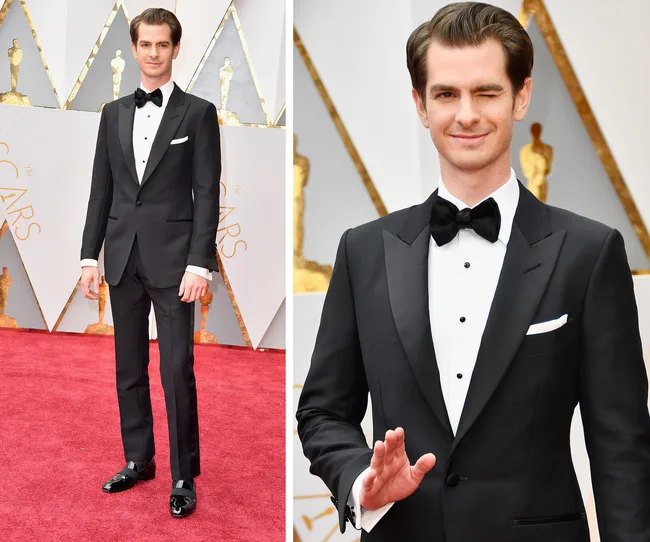 Andrew Garfield in a black tuxedo on the Oscars red carpet, smiling and winking at the camera.