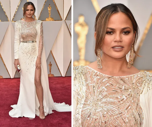 Woman in a white embellished gown on a red carpet with geometric backdrop, wearing long earrings.