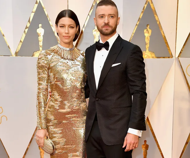 A couple poses on a red carpet event, the woman in a gold dress and the man in a tuxedo with a bow tie.