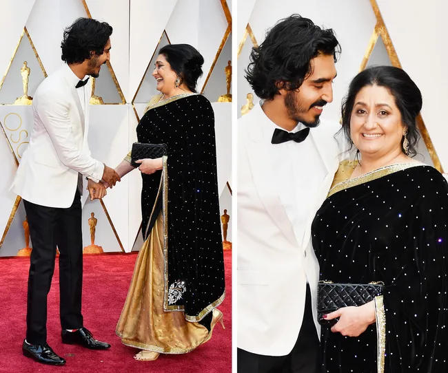Man in white tuxedo shaking hands and posing with woman in black and gold sari on red carpet at an event.