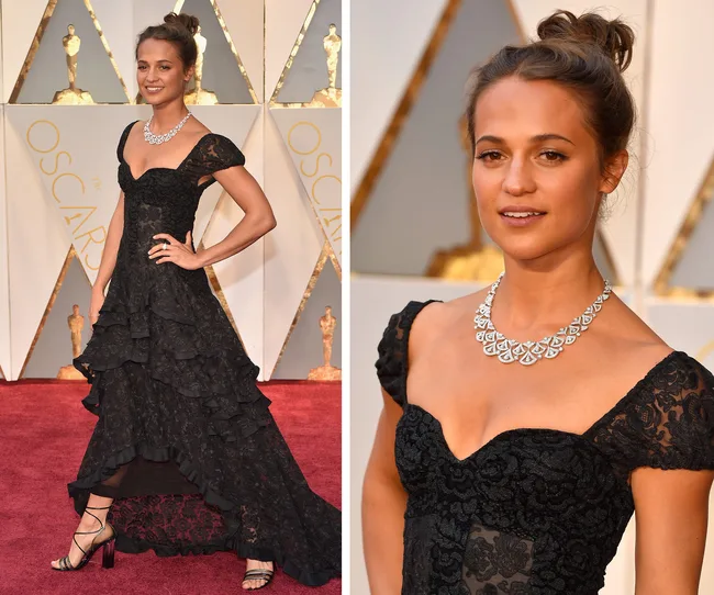 A woman in a black lace gown with a diamond necklace poses on the red carpet at an awards event.