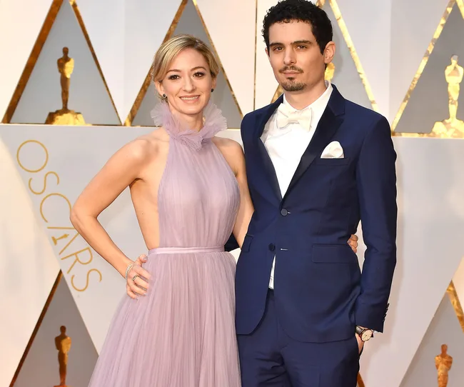 Two people pose at the Oscars; the woman wears a lavender gown, and the man wears a navy tuxedo with a white bow tie.