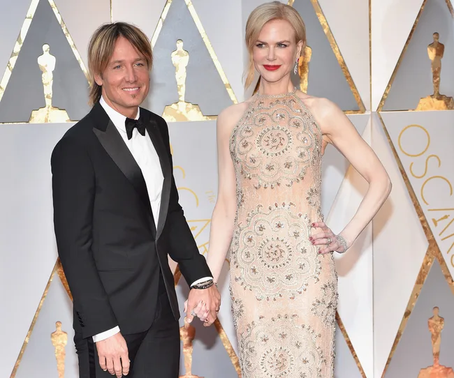 A couple in formal attire, holding hands on a red carpet with Oscar statuette backdrop.