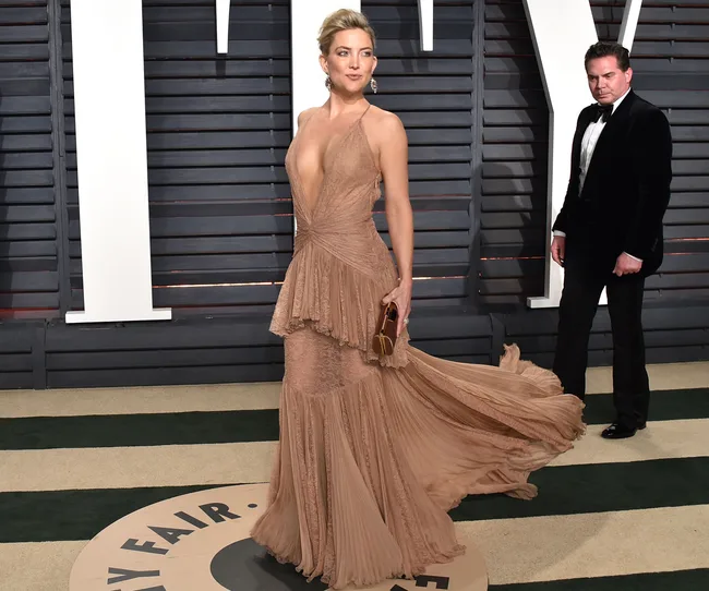 A woman in a flowing beige gown poses on a red carpet at a Vanity Fair event, while a man in a tuxedo stands nearby.