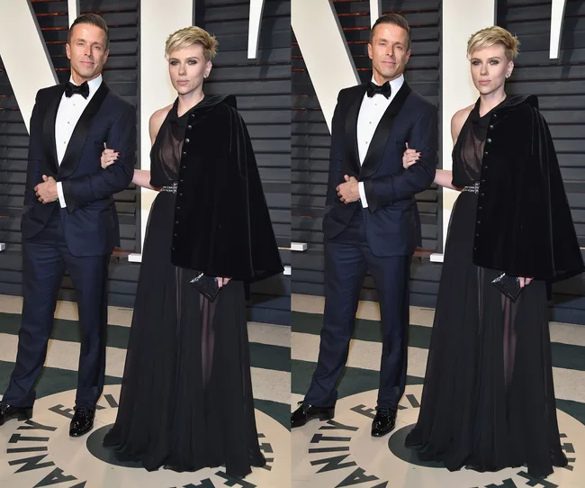 Couple posing on red carpet at an event; man in tuxedo, woman in black gown with a sheer overlay and cape.