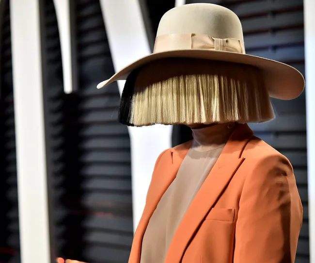 Person in orange suit, wearing a large hat and wig, covering their face completely.
