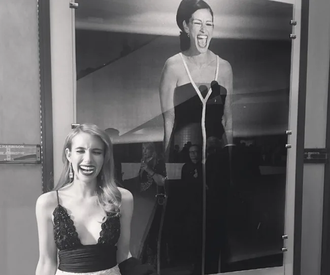 Woman smiling in front of a large photo of another woman laughing passionately in a formal gown, in black and white.