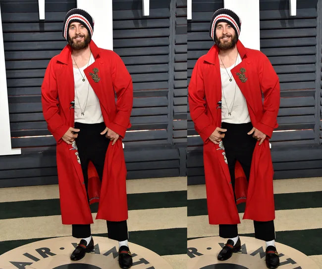 A person wearing a red coat, black pants, and a striped beanie, standing on a carpet with "Vanity Fair" written.